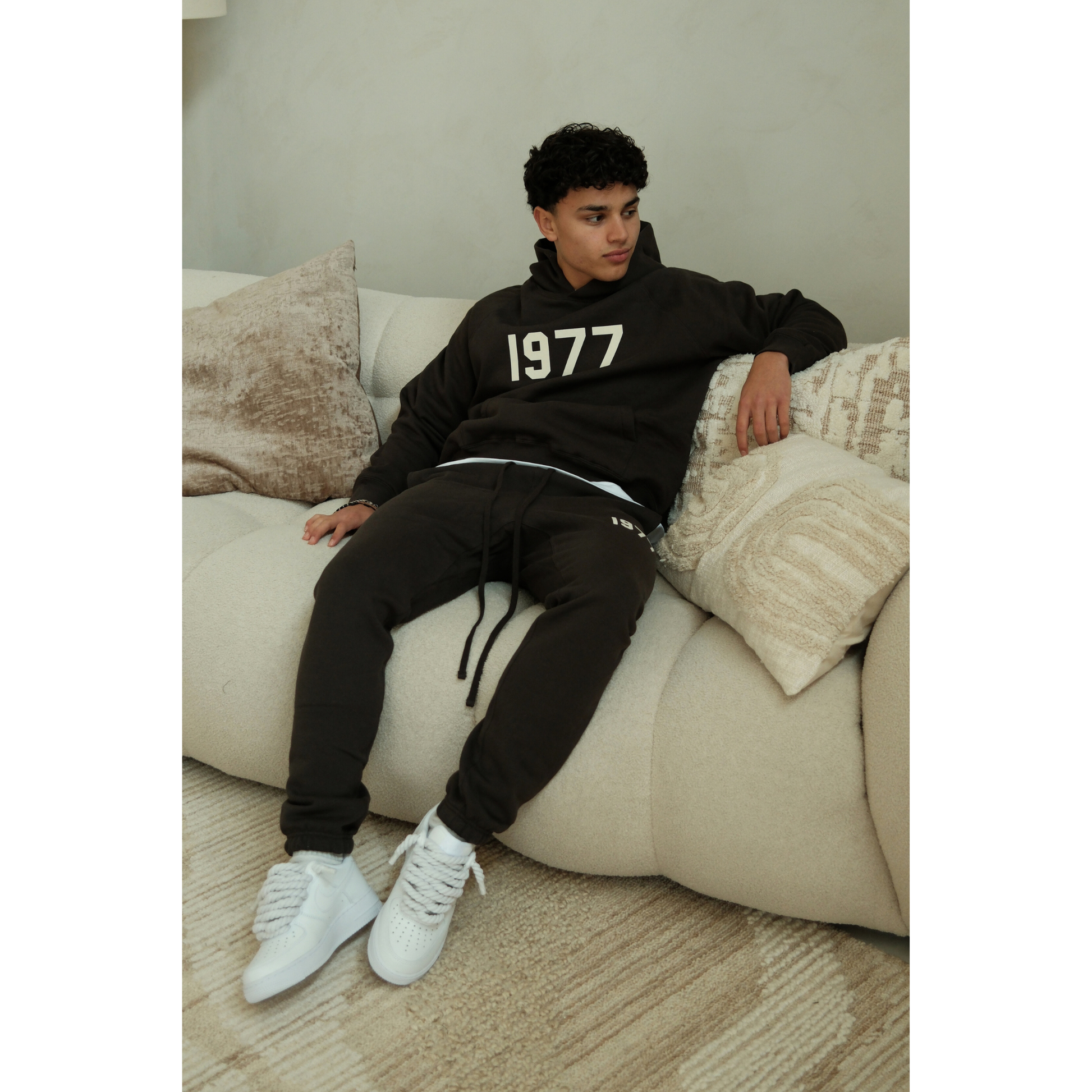 Fear of God Essentials 1977 hoodie and sweatpants set in iron black worn by model seated view